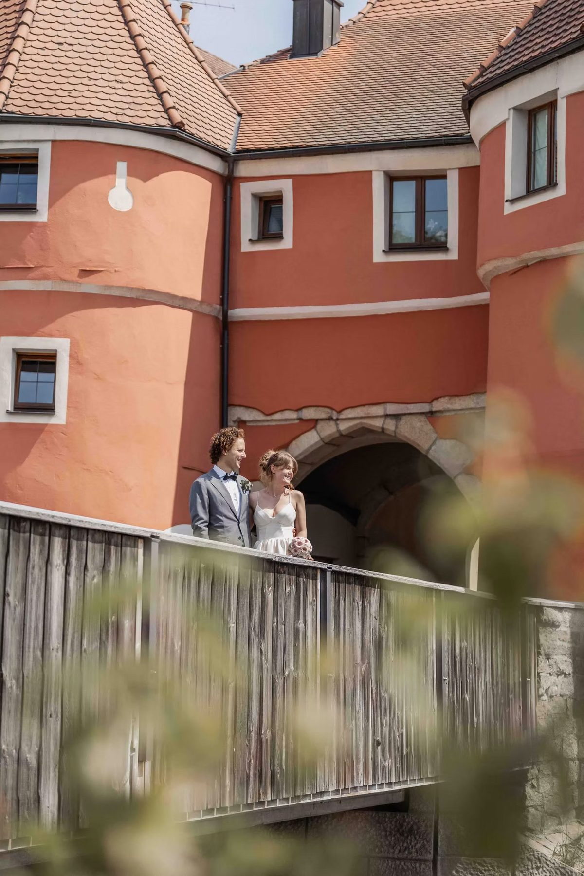 Brautpaar steht auf einer Holzbrücke vor einem roten Wasserschloss
